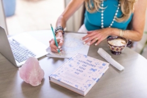 pink crystal, a book called "the astrology of you and me" and an astrology chart on the table with cynthia delaney